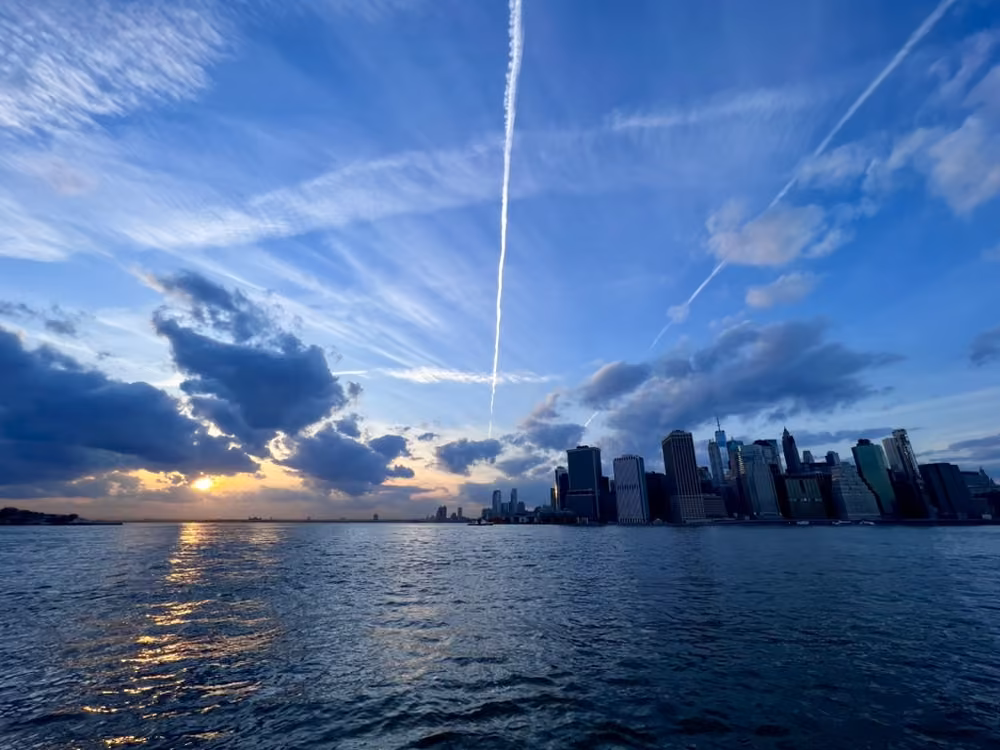 A city skyline across the water at sunset, under criss-crossing contrails